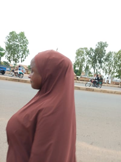 A medium shot captures a young woman from the side, her face obscured by the dark maroon hijab she wears, which drapes over her head and shoulders. She appears to be walking or standing near a paved road. In the background, the scene unfolds with a few key elements.
On the road, a motorcycle carrying two individuals is visible, moving away from the camera. The person in front is wearing a light-colored shirt and dark pants, while the passenger behind them is dressed in dark clothing. To the left of the motorcycle, a blue truck with a trailer is also on the road, though less prominently featured.
The setting appears to be an outdoor environment with a clear, overcast sky suggesting a daytime scene, likely in the morning or afternoon given the illumination. Trees with green foliage line the background, indicating a somewhat rural or suburban area, and some buildings are partially visible behind the trees. A concrete barrier separates the road from the area where the trees and buildings are located. The overall impression is a snapshot of daily life in Tumfure, Nigeria, with everyday transportation and a common scene of people on the move. There is no visible text in the image.