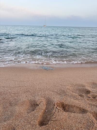 A wide shot of the beach at Blanes, Spain, captures a serene seascape at dusk. The calm, blue ocean stretches to the horizon, dotted with a solitary sailboat. Gentle waves lap at the shore, creating a foamy edge along the wet sand. The beach itself is covered in fine, light brown sand, with footprints clearly visible in the foreground, suggesting recent human presence. A single, dark, rectangular object, possibly a rock or debris, rests on the wet sand near the waterline. The sky above is a pale, hazy blue, blending into a soft lavender hue at the horizon, indicating the transition from day to night. The scene evokes a sense of peaceful solitude, characteristic of a quiet evening by the sea.