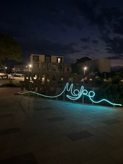 The image shows a nighttime scene in an urban setting, likely in Shchelkino, Ukraine. In the foreground, a bright blue neon light sculpture is prominently displayed, forming an abstract shape with flowing lines. This sculpture is situated on a paved walkway, characterized by rectangular stone tiles.
Behind the neon sculpture, a large, three-dimensional wooden sign with carved letters is visible. The letters are thick and appear to spell out a word or phrase, though the full text is not entirely legible due to the angle and lighting. Some of the letters appear to be "ТОР", and potentially other letters that are less clear.
The background reveals a multi-story building with windows, illuminated from within, suggesting it is occupied or lit. Beyond the building, trees and other structures are visible, some silhouetted against the dark night sky. The sky itself is filled with clouds, adding a dramatic element to the scene. Streetlights are also present, casting pools of light on the ground and illuminating parts of the environment.
The overall impression is one of a public space at night, possibly a plaza or park area, adorned with artistic installations. The time of day is clearly night, and the cloudy sky suggests there might be no moon or that it is obscured. The presence of buildings and streetlights confirms an urban environment. No specific event appears to be taking place; it seems to be a static display of art and urban architecture.