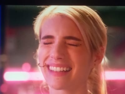 The media file is a close-up, low-resolution still image of a young Caucasian woman with blonde hair. She is smiling with her eyes closed, her teeth visible. She appears to be outdoors, with blurred neon lights in the background, suggesting a nightlife or entertainment setting. The lighting is predominantly pink and purple, casting a glow on her face. There are no visible objects or text. The woman's expression conveys happiness or amusement. The time of day is likely evening or night due to the artificial lighting and blurred background. There is no information about the weather. The location context provided is Sofiivska Borshchahivka, Ukraine, but there are no visual cues within the image to confirm this location.