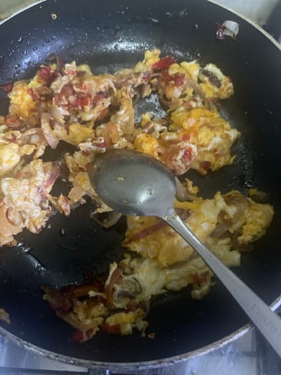 A top-down, close-up shot shows scrambled eggs with red onions and red peppers being cooked in a non-stick black frying pan on a stovetop. A stainless steel spoon rests in the pan amongst the eggs. The environment around the stove appears to be a kitchen setting. There is no indication of the specific location within Rafin Bauna, Nigeria. No people are visible.