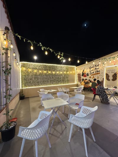 The image captures an outdoor night scene at a cafe or restaurant patio in Chennai, India, illuminated by string lights and wall lighting. The dark night sky, with a visible moon, sets a tranquil backdrop. White, lattice-patterned tables and chairs are arranged across a tiled floor, with some chairs stacked. A single person, dressed in a black and white patterned top and red pants, is seated at a table against a vibrant mural.
The mural, rich with cartoon-style depictions of people, food items like pizza and burgers, and musical notes, is adorned with text reading "Good Food Good Mood Good Vibes THAT'S ALL WE NEED". To the right of the mural, a prominent sign features angel wings and the text "MADRAS FOOL WALK" (likely "FOOD WALK"), along with instructions like "Be FIRST Selfie!", "Stand here Smile big Post often!". This suggests the location is designed for photo opportunities and a pleasant dining experience.
Along the roofline and a vertical wall section, decorative string lights with individual bulbs are intertwined with leafy garlands, casting a warm glow. The vertical wall also features a mesh-like pattern with integrated lighting, adding to the ambiance. A potted plant with white pebbles is visible near the foreground. The overall atmosphere is relaxed and inviting, indicative of an evening outing at a casual eatery.