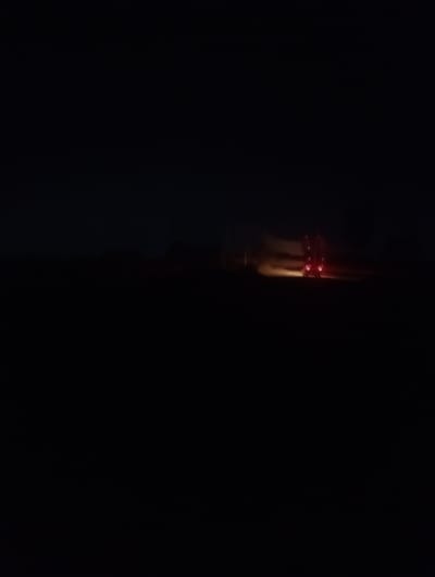 In the city of Reifild-Ist, Nigeria, a dark scene unfolds. A vehicle, possibly a truck, is seen from the rear, its red taillights illuminated against the blackness of night. The vehicle appears to be stationary or moving slowly, with a faint glow emanating from its headlights, casting a diffused light on the ground ahead. The surrounding environment is a vast expanse of darkness, suggesting an open area, perhaps a rural road or a sparsely populated region. The overall atmosphere is one of solitude and stillness, with the glowing taillights being the only discernible feature in the deep night.