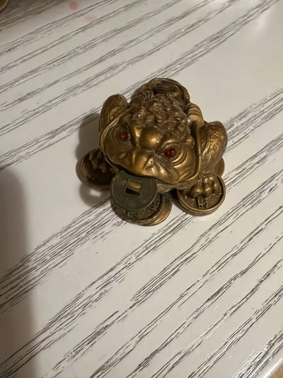 The image displays a close-up, overhead view of a brass figurine of a three-legged toad with red jewel eyes. The toad is positioned on a surface with a white background and diagonal dark lines, resembling wood grain or a patterned fabric. The figurine has a detailed, textured surface and appears to be a decorative item, possibly a feng shui charm. It is adorned with what seem to be Chinese coins, one prominently placed on its back and another near its head. The lighting suggests an indoor setting, possibly during the day, as there are no strong shadows indicative of direct sunlight. The overall mood is neutral, focusing on the object itself. No people are visible in the frame. The context of Athens, Greece, does not provide any specific visual cues within the image to support or contradict this setting, as the image focuses solely on the object and its immediate background. There is no visible text in the image.