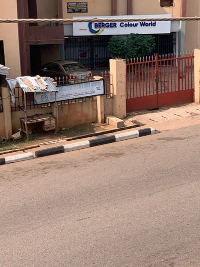 The image shows a street view in Enugu, Nigeria. In the background, there is a building with a sign that reads "BERGER Colour World". In front of the building, there is a red gate and some greenery. To the left, there is a small stall with a desk and a sign that says "ADVISORY COUNCIL OPAC". A car is parked in the driveway. The street in the foreground is paved and has a white and black painted curb.