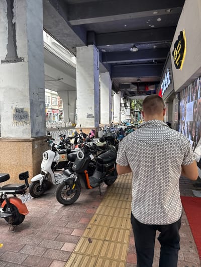 In Yuexiu District, China, a man with a short buzz cut is walking away from the camera. He is wearing a white short-sleeved shirt with a repeating black geometric pattern and black pants. The man is walking on a sidewalk paved with red and yellow textured tiles. To his left, a row of electric scooters and motorcycles are parked. Some of these vehicles are white, black, and orange. One of the white scooters has a license plate that reads "TV3219". To his right, a building features a wall covered with posters. Above the sidewalk, a concrete overpass structure is visible, with thick concrete pillars supporting it. The environment appears to be an urban area, possibly a commercial street, with buildings and infrastructure characteristic of a city. Graffiti is present on one of the concrete pillars.