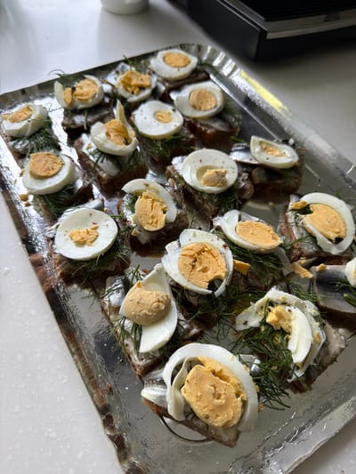 A tray of food, likely prepared for a gathering or meal, sits on a white surface. The tray is lined with crumpled aluminum foil, reflecting the overhead light. Arranged on the foil are numerous open-faced sandwiches. Each sandwich appears to be on a slice of dark bread, topped with small fish, likely sprats or anchovies, and garnished with fresh dill. Dominating the toppings are slices of hard-boiled eggs, cut in half or quarters, revealing their yellow yolks. Some of the egg pieces are slightly discolored, with hints of green or brown, suggesting they may have been seasoned or dressed. A few red pepper flakes are visible on some of the egg slices. The overall presentation suggests a home-prepared dish, possibly for a festive occasion or a light meal. The lighting is bright, indicating it might be daytime or under strong artificial lights. There is no visible text in the image.