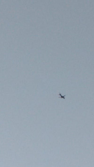 This media file shows a small airplane flying in a pale blue sky. The airplane is dark in color and appears to be a model or a toy, rather than a full-sized aircraft. It is positioned in the upper right quadrant of the frame and is slightly tilted. The sky is overcast or hazy, with no discernible clouds, suggesting it could be daytime but not necessarily clear weather. There are no other objects or features visible in the frame, creating a minimalist composition focused solely on the airplane against the sky. No text is visible in the image. The context provided indicates the location is Riverhead, USA.