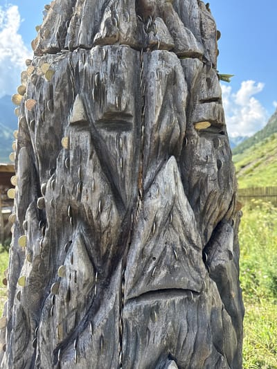 The image shows a close-up of a weathered, carved wooden totem-like structure in Chemal, Russia. The surface of the wood is textured with deep grooves and rough patches. Numerous coins of varying sizes and colors are embedded in or affixed to the surface of the structure, particularly in the crevices and around the carved features. The carvings appear to depict abstract facial features or symbols. In the background, blurred greenery and a blue sky with scattered clouds suggest an outdoor, possibly mountainous, environment. A wooden fence and some distant buildings are faintly visible. The focus is tightly on the wooden object and the coins.