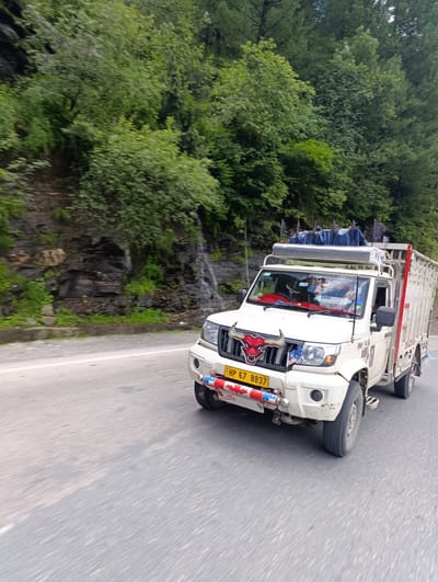 The image shows a white pickup truck driving on a road in Palchan, India. The truck is decorated with a bull's head emblem on the front grille and has several flags on its roof. The surrounding environment is lush green with trees and a rocky cliff face with a small waterfall. The road appears to be paved and has a white line marking the lanes. The truck is captured in motion, with a slight blur indicating its speed.