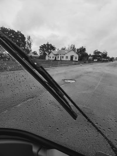 The black and white photograph presents a view from inside a vehicle, looking out through a rain-splashed windshield. Numerous water droplets cling to the glass, suggesting recent or ongoing rain. Two windshield wiper blades are prominently featured in the foreground, resting horizontally at the bottom of the frame, slightly angled upwards to the left. A portion of the vehicle's interior, possibly the right door frame or dashboard, is visible along the very right edge.

Beyond the wet glass, a wide, wet asphalt road or parking lot stretches across the mid-ground, showing reflective patches from the water. A distinct, light-colored puddle or pothole filled with water is visible on the road surface. In the background, on the left side of the scene, stands a simple, light-colored building, possibly a two-story house or institutional structure, characterized by several rectangular windows. Lush, dark trees with dense foliage are clustered around and behind the building. The sky above is overcast and bright, indicative of a cloudy day.

The scene captures a moment during or shortly after rain, evoking a quiet or subdued atmosphere. The time of day appears to be daytime, though the heavy clouds obscure any direct sunlight. No people are visible, and there is no discernible text in the image. The setting suggests a semi-urban or rural environment, consistent with the provided location context of Vom, Nigeria.