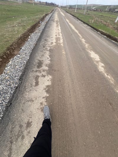 The image displays a rural road in Qoraobdol QFY, Uzbekistan, viewed from a first-person perspective looking forward. In the immediate foreground, the lower left quadrant shows the left leg and foot of an individual, wearing black pants and a grey athletic shoe, positioned on the left edge of the roadway. The road itself is partially constructed or undergoing repair; the leftmost lane consists of a recently applied layer of dark asphalt and light-colored gravel, bordered by an excavated dirt trench. The central and right portions of the road are unpaved, composed of packed earth and mud with numerous vehicle tire tracks and several wet, reflective patches. A white utility van or small bus is visible in the distant center of the road, appearing to move away from the viewer. Both sides of the road feature grassy fields, interspersed with utility poles extending into the distance. Low, rolling hills and scattered structures are discernible on the horizon under an overcast sky.