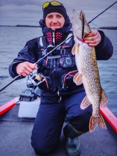 A Caucasian male, wearing a black beanie with yellow goggles, a black jacket with a red and black patterned collar, a grey life vest, and dark blue pants, kneels in a boat holding a large fish. The fish, likely a pike, is held by its mouth and gills, with its body extended. The man is smiling and looking at the camera. He is holding a fishing rod in his left hand. The boat appears to be on a body of water with a cloudy sky in the background. The location is identified as the city of Vysokoe, Russia.