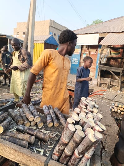 This image depicts a bustling street market scene in Gawuna, Nigeria, during daylight hours under a clear sky. The central figure is a man with dark skin and curly hair, wearing a long-sleeved orange tunic with small dark patterns. He stands with his right hand on a wooden display, looking towards the right side of the frame.
To his left, another man in a light yellow or cream-colored tunic, partially covered by a darker garment, is visible, smiling as he handles what appears to be sugarcane or wooden implements. Further to the right, a young boy in a blue traditional attire stands observing the man in orange. Other blurred figures can be seen in the background.
The foreground is dominated by large quantities of freshly cut sugarcane stalks, arranged in neat piles on two wooden display stands or crates. The sugarcane is dark brown-purple with pale, freshly cut ends. Wood shavings are scattered around the displays, suggesting recent preparation.
In the background, the setting includes simple market stalls or shops with corrugated iron roofs and walls, some painted in bright yellow and blue. A tall utility pole is prominent on the left. On a blue stall in the background, a partially visible sign reads, "GO FOR THE RICH CUSTARD." The overall atmosphere suggests a lively local commerce focused on the sale of sugarcane.