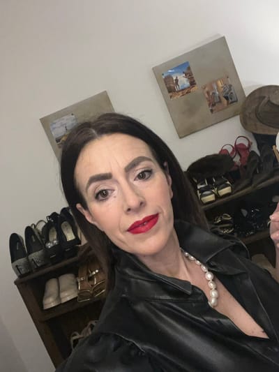 The image depicts a mid-adult female in an indoor residential setting located in the city of Konz, Germany. The female subject has long, dark brown hair parted in the middle, dark eyebrows, and is wearing red lipstick and dark eye makeup. She is wearing a black collared jacket, possibly leather or faux leather, and a large white pearl necklace around her neck. Her gaze is directed towards the camera, and her expression is neutral to slightly smiling.
The background consists of a light-colored wall. On the wall, two square-shaped framed items are visible. The item on the left contains a single image, while the item on the right contains two smaller images: an upper image depicting a person (male figure) against a structure, and a lower image showing multiple figures indoors. To the left of the subject, a wooden shelving unit functions as a shoe rack, holding numerous pairs of women's shoes of various styles and colors, including black heels with embellished rear portions and lighter-colored sandals. To the right of the subject, a dark brown wide-brimmed hat is partially visible, resting on an unseen surface. The lighting appears to be artificial.