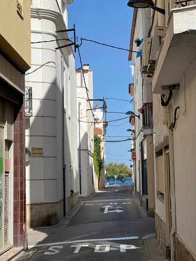 The image captures a narrow street in L'Escala, Spain, leading towards the Mediterranean Sea. On the left, a building with white walls and a textured facade displays a street sign that reads "CARRER SANT PERE." A black metal balcony railing is visible above the sign. Electrical wires crisscross the sky, supported by various poles and building mounts. On the right, buildings with white facades line the street, with visible air conditioning units mounted on one. The street surface is asphalt, marked with white directional arrows and the word "STOP" painted in large white letters. In the distance, a tree stands near the blue sea, with a person seated by the water. Two red stop signs are posted on the right side of the street. The sky is a clear, bright blue.