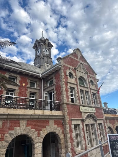 The image shows the Muizenberg Railway Station building in Muizenberg, South Africa. The structure is primarily built of red brick and grey stone, with a distinctive clock tower visible against a backdrop of a cloudy blue sky. The building has multiple stories, with windows and architectural details such as arches and stone trim. A sign bearing the name "Muizenberg Station" is affixed to one side of the building. Additional signage is partially visible on the right side of the image, possibly indicating a restaurant or cafe within the station. A metal railing is present on a balcony. Palm trees are visible at the bottom of the frame.