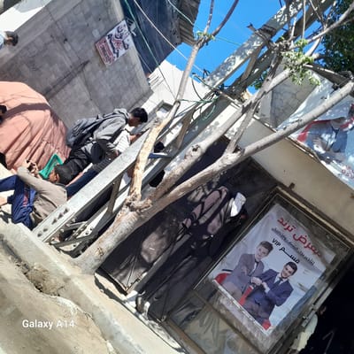 A sideways-angled shot taken in Khan Yunis, Unknown, shows several individuals in a cluttered urban scene. One person is seated on a metal structure, holding a mobile phone. Another individual with a backpack is standing, possibly interacting with something near a building. A brown fabric covers a section nearby. A tree branch obscures part of a shopfront with a poster featuring two young men in suits. Another poster is visible higher up on the building. A suit is hung over the shopfront. Power lines are visible overhead. The environment suggests a busy, possibly impoverished, urban area.