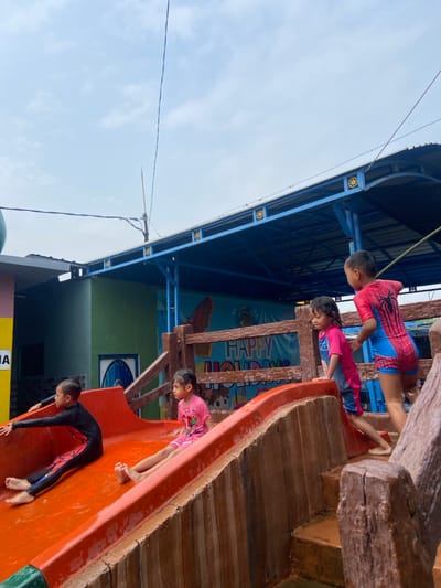 The image shows four children playing on a water slide at a waterpark in Kecamatan Tanggulangin, Indonesia, located at coordinates -7.497 latitude and 112.684 longitude. One child is sitting on the slide, another child is climbing the steps to the slide, and two children are standing by the slide. The slide is orange and red and is made of plastic. The steps are made of wood. There is a wooden fence behind the slide with a sign that says "Happy Holidays." The children are wearing swimwear. The sky is cloudy and the weather is overcast. The ground is wet.