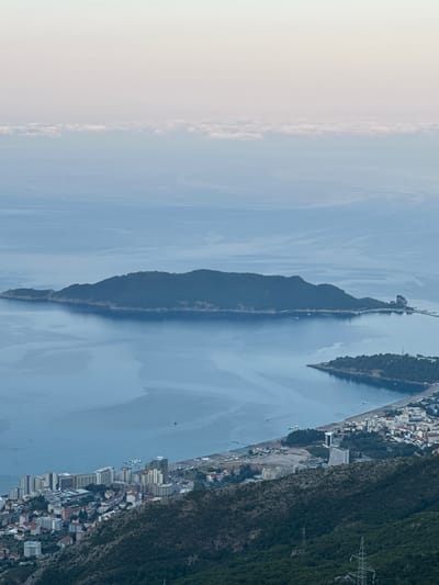 The image provides an elevated view of a coastal landscape, likely captured at dawn or dusk due to the soft, diffused light and pastel hues in the sky. In the distance, a large, elongated island covered in dense green foliage dominates the horizon, its shores meeting the calm, blue water. A narrow causeway or bridge appears to connect a portion of the island to the mainland, with a rocky outcrop visible at its eastern end.
Closer to the viewer, a smaller, tree-covered island juts out into the sea, forming a bay. Along the coastline, a strip of beach lined with numerous rows of sun loungers is visible, suggesting a resort area. Beyond the beach, a densely populated town unfolds, characterized by a mix of low-rise buildings with red-tiled roofs and taller, more modern structures, including hotels and apartment complexes. The town extends inland, appearing to climb the slopes of the hills that frame the scene.
The foreground is dominated by a steep, densely forested hillside. A prominent metal electricity pylon stands on the slope, indicating infrastructure within the natural landscape. The sea itself is a tranquil expanse of blue, with subtle ripples and reflections indicating a calm surface. A few small boats are scattered across the water, one near the town and others closer to the islands. The sky above is a gradient of pale pink, lavender, and soft blue, with a distinct band of white clouds stretched across the horizon. The scene depicts a coastal settlement nestled between the sea and mountainous terrain, with islands providing a dramatic backdrop. Based on the geographical features and architectural style, this location is likely Brajici, Montenegro.