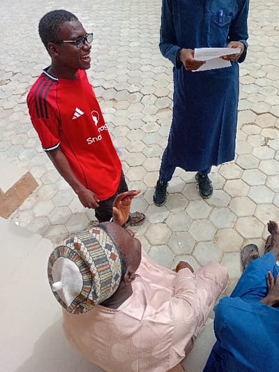 The scene, located in Kwazailewa, Nigeria, depicts an outdoor gathering of at least four individuals on a paved surface composed of light-colored hexagonal pavers and a lighter, possibly tiled, area in the lower left. The lighting indicates daylight.
In the foreground, an older male is seated directly on the paved ground, wearing a light peach-colored patterned long-sleeved garment and a multi-colored patterned cap with a white central dome. He gestures with his raised right hand, palm open, and looks upwards towards the standing individuals. To his right, partially visible, is another seated individual wearing blue trousers and dark slip-on shoes.
Standing to the left, a male in dark-framed glasses and a red Adidas t-shirt with black shoulder stripes is smiling broadly and looking towards the seated older male. He wears brown sandals.
To the right, another individual stands, wearing a dark blue traditional long garment over dark trousers and black athletic shoes. This individual holds a folded white document in both hands at waist level; a blue pen is visible in their chest pocket. The head of the individual in the dark blue garment is not visible.
The interaction suggests an ongoing discussion or address among the group.