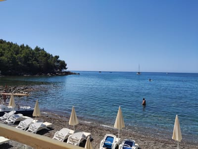 The image depicts a scenic beach in Zagrađe, Montenegro. In the foreground, several empty white sun loungers are arranged on a pebble beach, some covered by closed beige umbrellas. A small white pedal boat with the number '5' visible on its side is partially submerged at the water's edge, accompanied by a wooden sunshade. To the left, a rocky shoreline is covered with dense green pine trees.
The calm, clear blue sea stretches out to the horizon under a bright, cloudless sky. Several small boats and a sailboat are visible in the distance. A few people are in the water; one person is standing knee-deep in the sea on the right side of the frame, and another individual is on a paddleboard further out. The water is transparent, revealing the pebbles and rocks beneath the surface near the shore. The overall impression is one of a tranquil and sunny day at the beach.