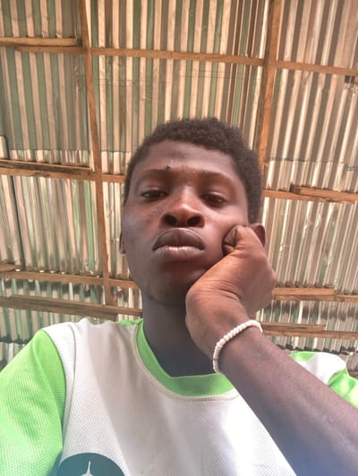 A dark-skinned male, located in Agege, Nigeria, is shown from the chest up, looking directly at the camera. He has short, dark, curly hair and a small mole superior to his right eye. His right hand is positioned with the palm against his chin. A white beaded bracelet is present on his left wrist. He is wearing a white t-shirt with light green trim on the collar and shoulders. The background consists of corrugated metal sheets, likely a roof or wall, supported by horizontal and vertical wooden beams.