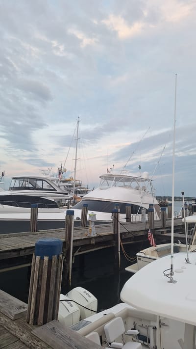 The image depicts a marina in Nantucket, United States of America, with multiple yachts docked. In the foreground, a wooden dock runs horizontally, with several pilings, one capped with a blue object, supporting it. Two outboard motors, branded "Mercury," are visible attached to the stern of a white boat. Beyond the foreground, another wooden dock is present, with a white yacht docked to it, featuring fishing rods extending from its superstructure. To the left, a larger white yacht with a dark hull and enclosed cabin is docked. In the background, more yachts and a sailboat are visible. The sky is overcast with soft clouds, suggesting either early morning or late evening. The overall scene is one of a tranquil marina setting.