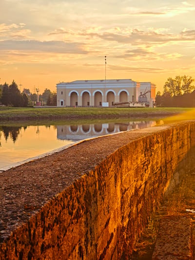 The image captures a sunrise scene at a waterfront, likely in the city of Vodnik SNT, Russia. In the foreground, a weathered stone wall, illuminated by the warm, golden light of the rising sun, runs diagonally across the frame. The wall's texture is rough, with visible moss and irregularities. Beyond the wall, a calm body of water reflects the sky and the structures on the opposite bank. The water's surface is smooth, with subtle ripples. Across the water, a long, light-colored building with a series of arched openings stands prominently. This building is set against a backdrop of greenery, including trees and well-maintained grass. The sky above is painted with soft, warm hues of orange and yellow, with scattered clouds catching the sunlight. Electrical poles and wires are visible in the distance, suggesting the presence of infrastructure. A small flag is atop a pole in front of the building. The overall atmosphere is serene and tranquil, bathed in the soft glow of dawn. There are no people or animals visible in the frame.