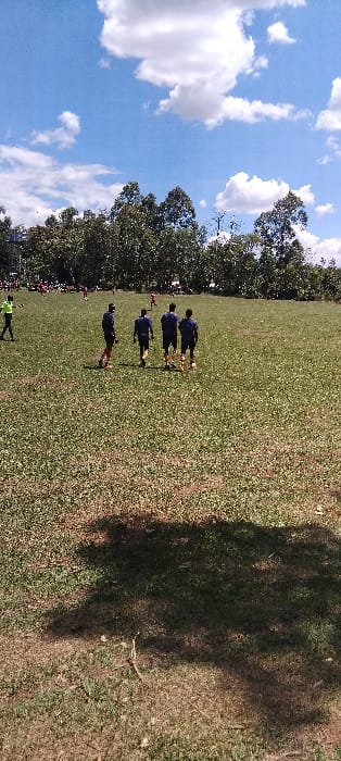 The image shows a group of four men wearing blue shirts walking across a grassy field in Bungoma, Kenya, located at 0.554 latitude and 34.774 longitude. The men are all facing away from the camera and appear to be walking in a line. The field is covered in green grass and has a large, shadowy tree in the background. The sky is blue and partly cloudy. There is a line of trees on the far edge of the field. There are several people visible in the distance near the edge of the trees.