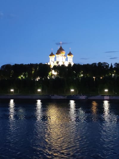 The image depicts a wide shot of a body of water reflecting lights from a cityscape at dusk. In the background, a large, white building with golden domes and crosses is illuminated, possibly a church or cathedral. It is situated behind a dense line of dark green trees. Along the waterfront, several lampposts are visible, casting pools of light on the ground. The water in the foreground shows strong reflections of the illuminated buildings and lampposts, creating streaks of yellow and white light on the dark blue surface. The sky is a deep, clear blue, indicating the transition from day to night. There is no indication of the specific location being in Шебунино, Russia, or any specific city.
