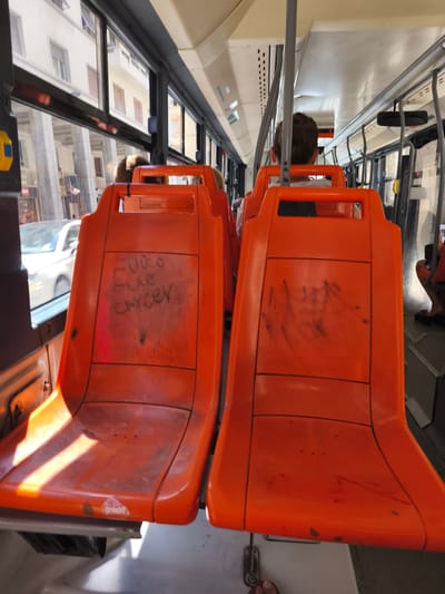 The image shows the interior of a bus in Livorno, Italy. Two bright orange plastic seats are prominently featured in the foreground, facing forward. The seat on the left has graffiti scribbled in black marker on its backrest, which appears to read "FUORO alle cancer". The seat on the right has some smudged black markings on its backrest. Sunlight streams in through the windows, casting a bright diagonal stripe across the seat on the left. Through the windows, buildings and street activity are visible. In the background, other passengers are seated, and the ceiling of the bus is visible. The overall impression is that of a public transport vehicle with some signs of wear and vandalism.