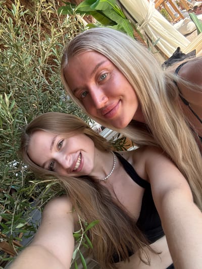 Two women are taking a selfie outdoors, with lush greenery in the foreground and beach chairs and canopies in the background, suggesting a resort or beachside location. The woman in the foreground, on the left, has long brown hair and is smiling, wearing a black top and a pearl necklace. The woman behind her, on the right, has long blonde hair and is also smiling. The scene is brightly lit, indicative of daytime. The location is identified as Drobnići, Montenegro.