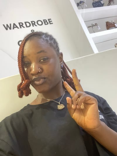 A young Black woman with braided auburn hair is posing for a selfie indoors. She is wearing a black t-shirt and a silver necklace with a pendant. She has a nose ring and a small piercing above her lip. Her left hand is raised, with two fingers extended in a peace sign. The background shows a wall with the word "WARDROBE" printed on it, and shelves stocked with handbags. The scene appears to be a retail store or a boutique.
