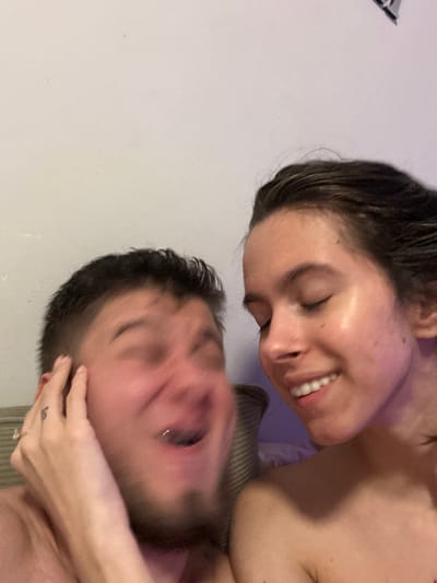 Two individuals, an adult male and an adult female, are captured in a close-up indoor photograph within a residence in Corby, United Kingdom. The background consists of a plain, light-colored wall, with a striped textile object partially visible behind the male's left shoulder. The female, positioned on the right, has dark hair, which appears damp, and is smiling with her eyes closed. Her bare right shoulder and upper chest are visible, illuminated by a faint purple light from the right. The male, on the left, has short dark hair and facial stubble. His face is motion-blurred, suggesting movement, with his mouth open and a lip piercing visible. A hand, presumed to be the female's, is placed on the side of the male's head; a small black tattoo is present on her index finger, and a ring on her ring finger. Both subjects appear to be shirtless or in minimal attire.