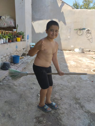A young boy, appearing to be of Central Asian descent, stands in a sunlit outdoor courtyard in Yangibazar, Uzbekistan. He is shirtless, wearing black shorts and blue sandals. The boy is holding a broom horizontally with both hands and giving a thumbs-up with his right hand, smiling broadly at the camera. The broom has a light-colored wooden handle and a green and white bristled head.

The courtyard has a dirt and gravel ground. To the left of the boy, a low wall with bricks and a concrete top runs along a building. On this wall, several potted plants are visible, including a bright yellow pot and a white pot with red flowers. Behind the plants, a white sheet is draped over something that resembles a bed. Next to the wall, a blue bucket and a black bucket with a lid are placed on the ground, with a broom leaning against the wall near the blue bucket.

The background features a plain, light-colored wall with some graffiti and a coiled white rope hanging from it. The wall casts a long shadow of the boy onto the ground, indicating it is likely mid-afternoon. The sky is clear and blue, suggesting fair weather. In the distance, the top of some trees can be seen. The overall scene depicts a casual, everyday moment in a rural or semi-rural setting. No visible text is present in the image.