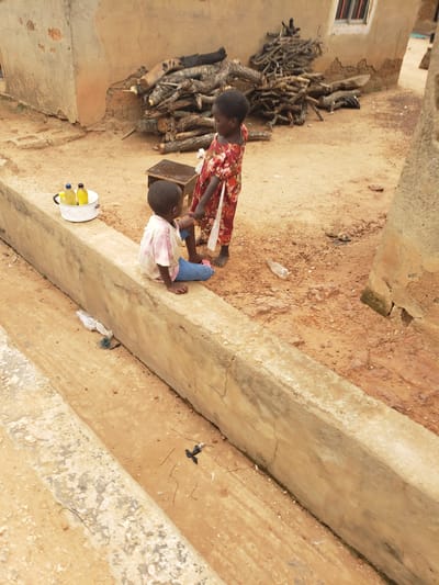 In a dusty, outdoor setting in Kachia, Nigeria, two young children are present. One child, facing away from the camera, sits on a concrete ledge. This child is wearing a light-colored, possibly tie-dye, t-shirt and blue pants. The other child, slightly older and also facing away, stands behind the first. This child is wearing a red dress with a floral pattern. The environment consists of a simple, earth-toned building with peeling plaster and a pile of firewood stacked against its wall. Two plastic bottles, one yellow and one clear with a yellow cap, are placed in a white pot with a handle on the ledge. There is a small wooden stool near the standing child. The ground is composed of dry soil and pebbles.