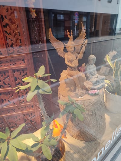 The image shows a shop window display in Rosenheim, Germany. The window is reflective, obscuring some details of the outdoor environment. Inside the window, there are several decorative items. Prominently featured is a carved wooden sculpture of a winged figure playing a stringed instrument, seated on a rock-like base. To the right of this sculpture is a smaller, stone statue of the Buddha in a meditative pose. In the foreground, there is a potted plant with green leaves and thorny stems. Further to the right, a tall plant with striped leaves is visible in a white pot. In the background, a carved wooden screen is partially visible. The overall scene suggests an establishment selling Asian-inspired decor or goods.