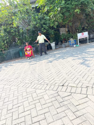 In Kalisampurno, Indonesia, a woman in a vibrant red and yellow dress sits on a chair outdoors, holding a bowl of food. Nearby, a man in a yellow polo shirt and dark pants stands, appearing to interact with her. The scene unfolds on a paved walkway with a herringbone brick pattern. Behind them, lush green trees and bamboo foliage form a dense backdrop. To the right, a white sign with red lettering, partially obscured, is posted on a stand. Various objects, including potted plants and trash bins, are visible in the background, suggesting a communal outdoor space. The lighting indicates it is daytime, with shadows cast by the foliage onto the ground.