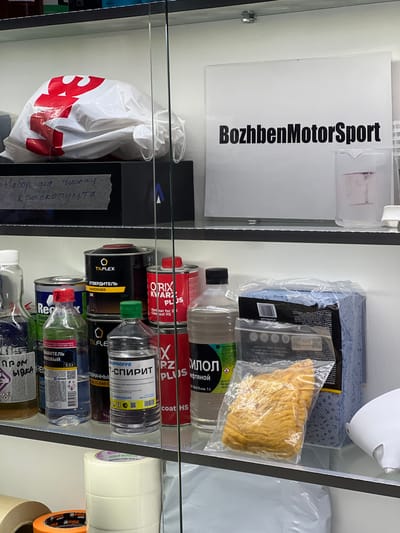 The image displays a retail shelf or display case stocked with various automotive care products, primarily related to car painting and cleaning. The items are arranged on multiple shelves, with some items in plastic packaging.

Prominently featured on the top left is a white sign with the black text "BozhbenMotorSport". Below this, a white plastic bag with a large red logo that appears to say "BMW" is hanging. To the right of the bag, on a grey strip of tape, handwritten Russian text reads "Набор для чистки краскопульта" which translates to "Set for cleaning spray guns".

The shelves below are filled with an assortment of bottles and cans containing liquids, likely solvents, cleaners, and paints. Several brands and product names are visible in Russian, including "TILFLEX", "ORIX KWAR", "REMIX", and "Plus". Some bottles have green and red caps. In the upper right, there are rolls of white and yellow tape, possibly masking tape or detailing tape.

The overall setting appears to be an automotive supply store or a workshop. The lighting is artificial, suggesting an indoor environment. There are no people visible in the image. The focus is entirely on the products displayed, indicating a commercial or organizational context for these items. The specific location "Veteran-2, Russia" provides geographical context for the origin or sale of these items.