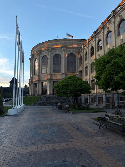 The image shows the Gyumri National Art Gallery in Gyumri, Armenia, at dusk. The building is made of light-colored stone and features classical architectural elements, including arched windows and ornate detailing. The Armenian flag flies atop the building. In front of the building is a plaza paved with cobblestones and a walkway lined with white flagpoles. Several benches are placed on either side of the walkway. Lush green trees and grass surround the building and plaza. The sky is a clear blue with hints of sunset colors. A dark, spire-like structure, possibly a church, is visible in the background to the left, partially obscured by trees. The scene is peaceful and well-maintained.