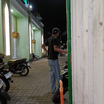 The image shows a man standing on a sidewalk in front of a building. The man is wearing a black shirt and blue jeans. He is looking to the right, possibly at a motorcycle that is partially visible in the image. The sidewalk is made of brick and has an orange cone placed in the middle of the sidewalk. The man is standing in front of a building with a green wall and a white roof. The building has windows on the side closest to the viewer. The building is lit by a light fixture on the front of the building, and the light fixture is illuminating the front of the building and the man in the image. There is a second motorcycle parked behind the man, and two people are visible seated on the sidewalk just behind the second motorcycle, possibly waiting for someone. The scene is located on the street in Kaliwungu, Indonesia (latitude: -6.961, longitude: 110.249).
