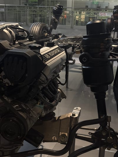 The image displays a V8 engine with "V8 FOUR CAM 32" text visible on its silver casing. The engine is mounted on a chassis, with a tire and suspension components also visible. The scene appears to be an exhibit within a museum or exhibition space, suggested by the polished floor and glass barrier in the foreground. In the background, there are industrial-style structures and some signage, possibly indicating a factory or a science museum. The overall lighting is artificial and somewhat dim, with reflections on the glass suggesting an indoor environment. Based on the context of automotive exhibits and the presence of such engines, this scene is likely located at the Toyota Commemorative Museum of Industry and Technology in Nagoya, Japan.