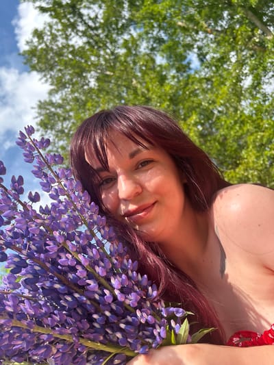 A woman with dark red hair is holding a bouquet of purple lupine flowers in front of her face. She is smiling and looking at the camera, with sunlight illuminating her face. Behind her, blurred green trees and a blue sky with white clouds are visible. The setting appears to be outdoors, likely in a natural environment.