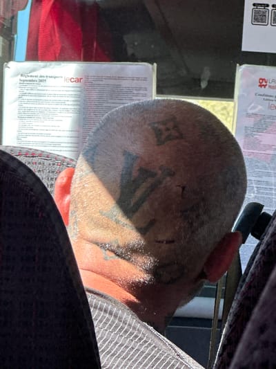 The image shows a man with a shaved head resting against a seat in what appears to be a bus or other public transportation vehicle. The man has several tattoos on his head, including what appears to be the Louis Vuitton logo, and other unidentified markings around his ear and temple. The bus windows feature posted notices. One partially visible poster on the window to the left mentions "Règlement des transports lecar Septembre 2025", indicating this is likely in France. The presence of "lecar" on the poster suggests the location may be Aix-en-Provence, France, as "lecar" is a bus service operating in that area. The lighting suggests the photo was taken during daylight hours. The man is wearing a striped shirt.