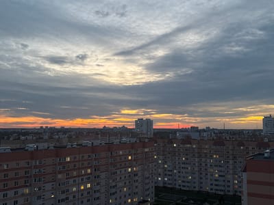 This is an aerial shot overlooking a city at dusk. The sky is a mix of grey clouds with streaks of orange and yellow from the setting sun. Below the sky are rows of apartment buildings, with many windows lit from within, suggesting it is evening. The buildings are modern and appear to be in a residential area of the city. In the distance, taller buildings can be seen, indicating a city landscape. The specific location is Homyel, Belarus.