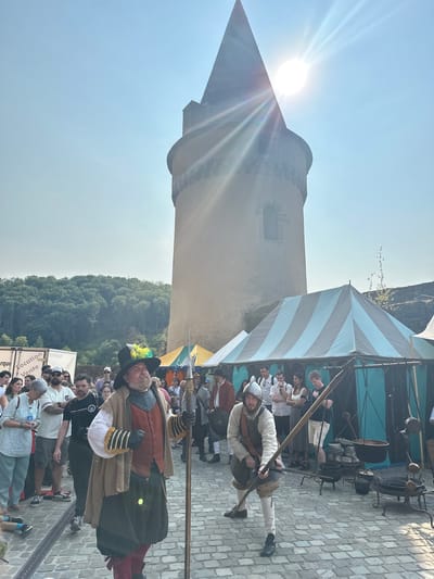 This image depicts a sunny outdoor scene at a historical reenactment event, likely held at or near the Vianden Castle in Vianden, Luxembourg. In the foreground, two men dressed in historical costumes are prominent. The man on the left wears a black hat adorned with feathers, a tan cloak over a red vest, and a green skirt-like garment. He holds a tall staff. To his right, another man in historical attire, wearing a helmet, a light-colored tunic, and knee-high stockings, holds a spear.

Behind them, a crowd of people in modern clothing are visible, suggesting they are spectators. Several tents with striped awnings are set up, one predominantly light blue and gray. In the background, a tall, cylindrical stone tower with a conical roof dominates the skyline. This structure, likely part of the Vianden Castle complex, is partially obscured by the bright sun which casts lens flares across the image. The ground is paved with cobblestones. The environment also includes a forested hillside in the far background and a clear blue sky with strong sunlight. Various historical-looking cooking vessels and equipment are displayed outside one of the tents. A trailer with French text that reads "Remorque Location Vente" is also visible on the left side of the frame.