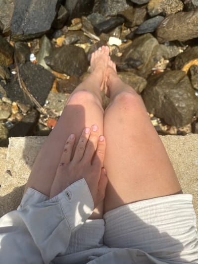 The image presents a top-down view of a person sitting at the edge of a body of water filled with rocks, likely in Walgama, Sri Lanka. The scene focuses on the person's legs and hands, with their bare feet partially in or resting near the shallow, clear water that reveals the rocky bottom. The person is wearing light-colored clothing, including what appears to be a light-colored, possibly ribbed, shirt and light shorts or skirt.

Bright sunlight illuminates the scene, casting distinct shadows across the person's legs, hands, and clothing, indicating a clear, sunny day, likely during the morning or afternoon. The person's left hand rests on their thigh, revealing a small, faded tattoo of three dots on the back of the hand and some minor scuffs or marks on the knuckles. Similar slight marks are visible on the knees. The individual is seated on a light-colored, flat concrete or stone surface that borders the rocky water. The overall impression is one of relaxation and connection with a natural, serene environment. There is no visible text in the image.