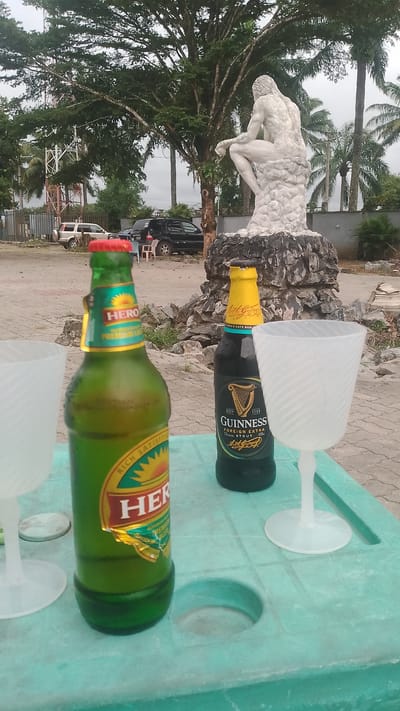 Two bottles of beer and two plastic wine glasses are placed on a green plastic table. One bottle is a green bottle labeled "HERO PREMIUM LAGER" with a red cap. The other bottle is a dark bottle labeled "GUINNESS FOREIGN EXTRA STOUT" with a yellow cap. The scene appears to be outdoors, with a statue of a man in a pensive pose in the background, surrounded by rocks and vegetation. Trees and palm trees are visible, suggesting a tropical or subtropical climate. In the background, there are vehicles, including a white SUV and a black car, and a fence. The sky is overcast, indicating a cloudy day. The overall mood suggests a relaxed, perhaps celebratory, outdoor gathering. The visible text includes brand names of the beers and their descriptions. The location context is Agbarho, Nigeria.