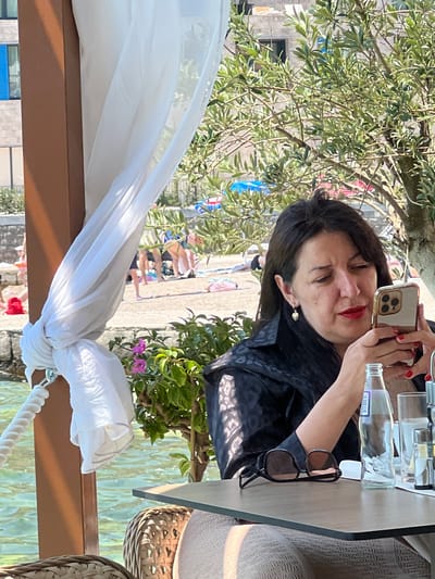 In Risan, Montenegro, a woman with dark hair and red lipstick is seated at a table, holding a smartphone in her right hand, and looking at its screen. She is wearing a black patterned jacket. On the table in front of her are a pair of sunglasses, a glass bottle of water, a glass, and other tableware. A white sheer curtain is draped on a brown wooden post to her left. Behind her, out of focus, is a beach with several people, some lying on towels and others standing. Sunlight filters through the leaves of a tree in the foreground, casting dappled shadows. The background shows buildings and a body of water.